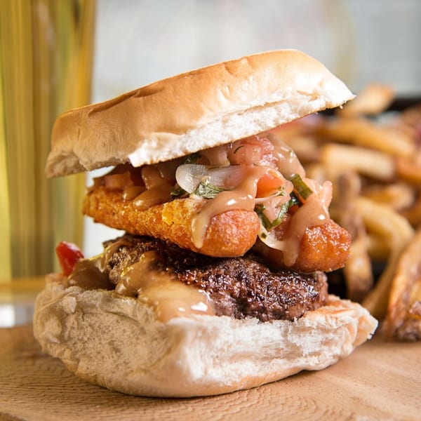 A gourmet burger with a thick patty, fried cheese, and relish, served with fries and a drink.