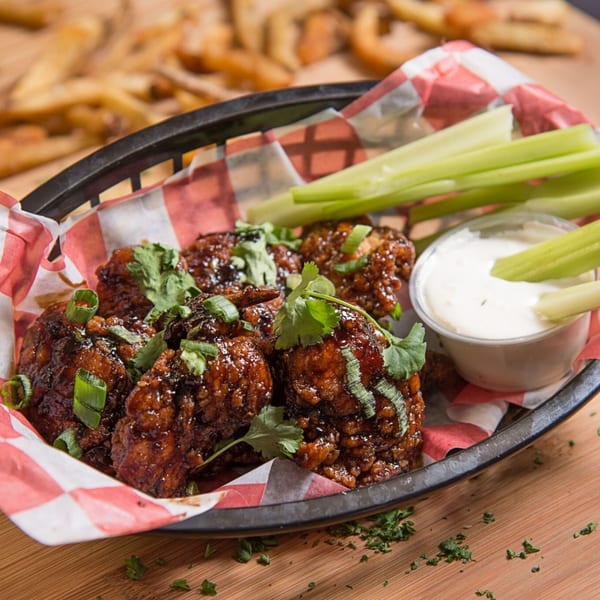 A basket of glazed chicken wings with celery sticks and ranch dressing, with fries in the background.