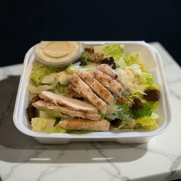 Chicken Caesar salad in a white takeout container with dressing on a marble surface.