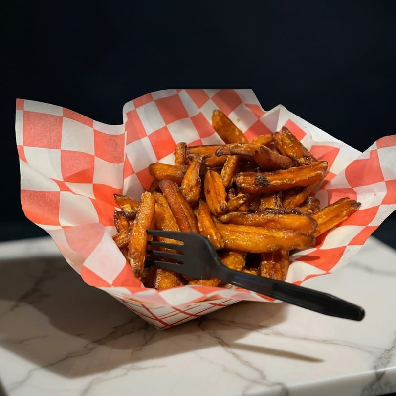 Sweet potato fries in a red and white checkered paper basket with a black fork.