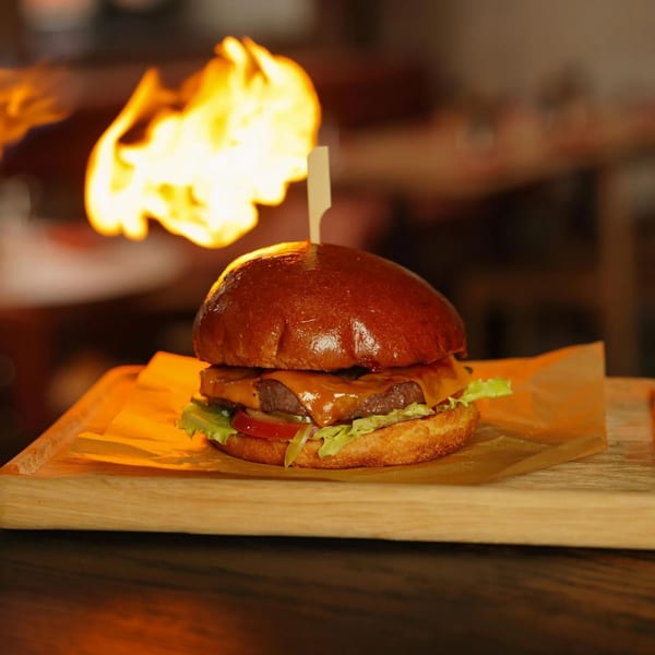 Gourmet burger with melted cheese, lettuce, and tomato, on a wooden board with flames in the background.